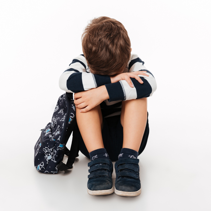 Un petit garçon triste est assis par terre la tête dans ses mains avec son sac à dos à côté de lui